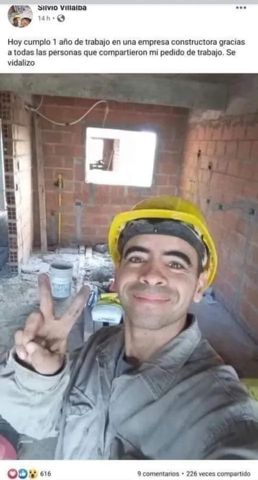 Un hombre con casco de construcción y ropa de trabajo se toma una foto haciendo el signo de la paz con la mano. Está en una habitación a medio construir con una ventana al fondo.